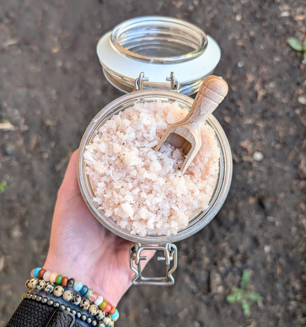 Gardener's Hand Scrub A hand holding a jar full of pink Gardener's Hand Scrub with a wooden scoop in it.