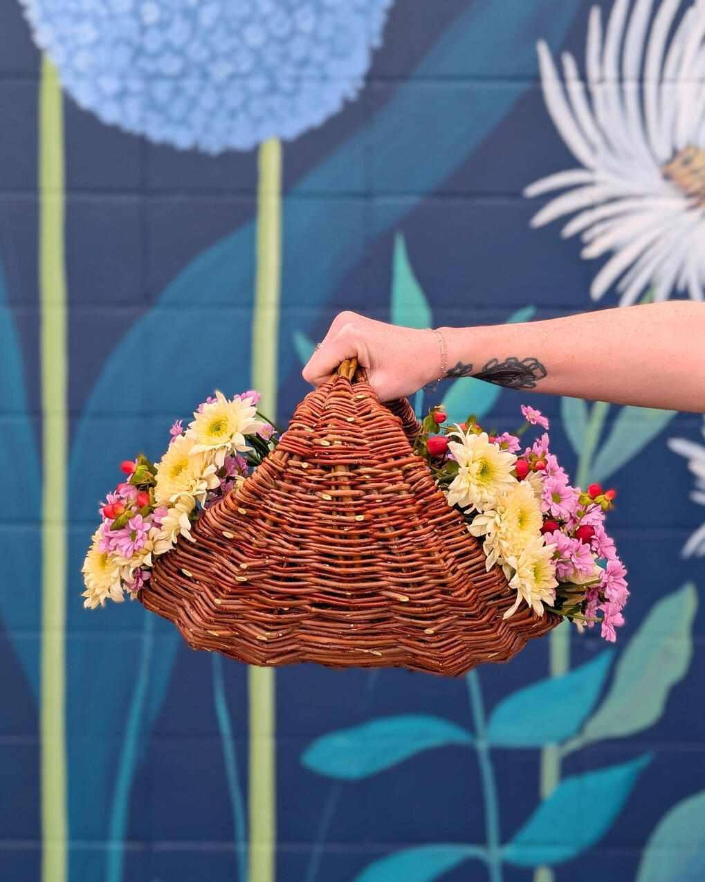 Gathering Basket A hand holding a basket filled with flowers, against a blue wall.